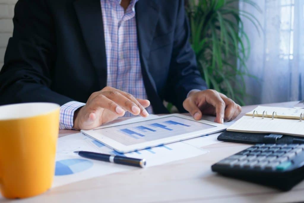 A businessperson interacts with a tablet displaying charts, with coffee and a calculator nearby. By Towfiqu barbhuiya on Unsplash.